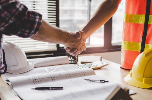 A man in a plaid shirt and a man wearing a construction vest shake hands over a desk. 